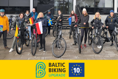 Representanter från partners i projektet Baltic Biking UPGRADE står framför en byggnad med sina cyklar och landsflaggor.