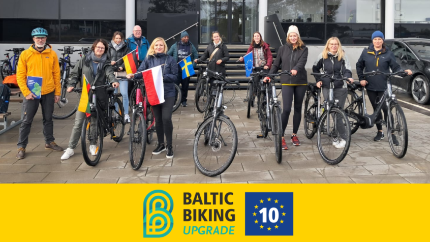 Representanter från partners i projektet Baltic Biking UPGRADE står framför en byggnad med sina cyklar och landsflaggor.