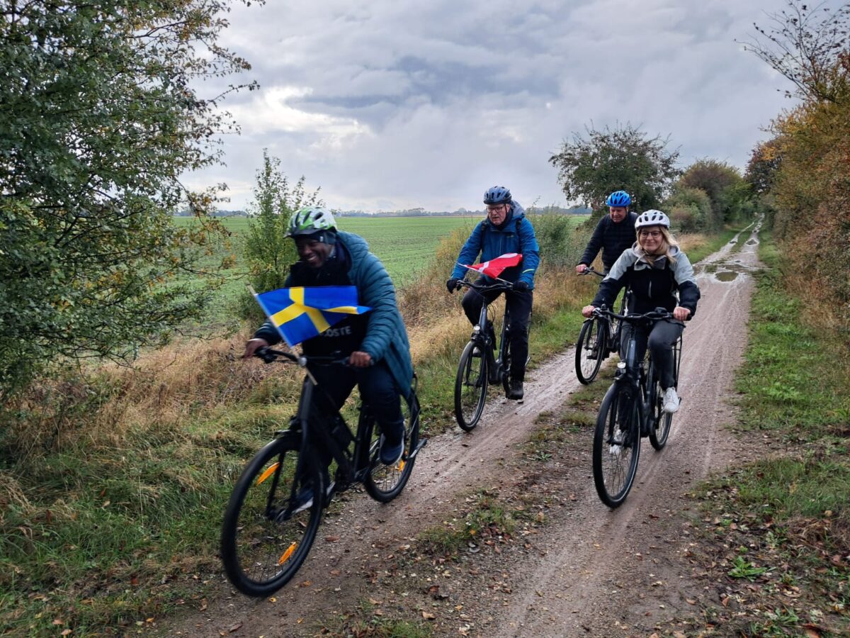 Fyra representanter för projektpartners i Baltic Biking UPGRADE cyklar i naturen med svenska och danska flaggan. Fotograf Tara Jacobsdotter.